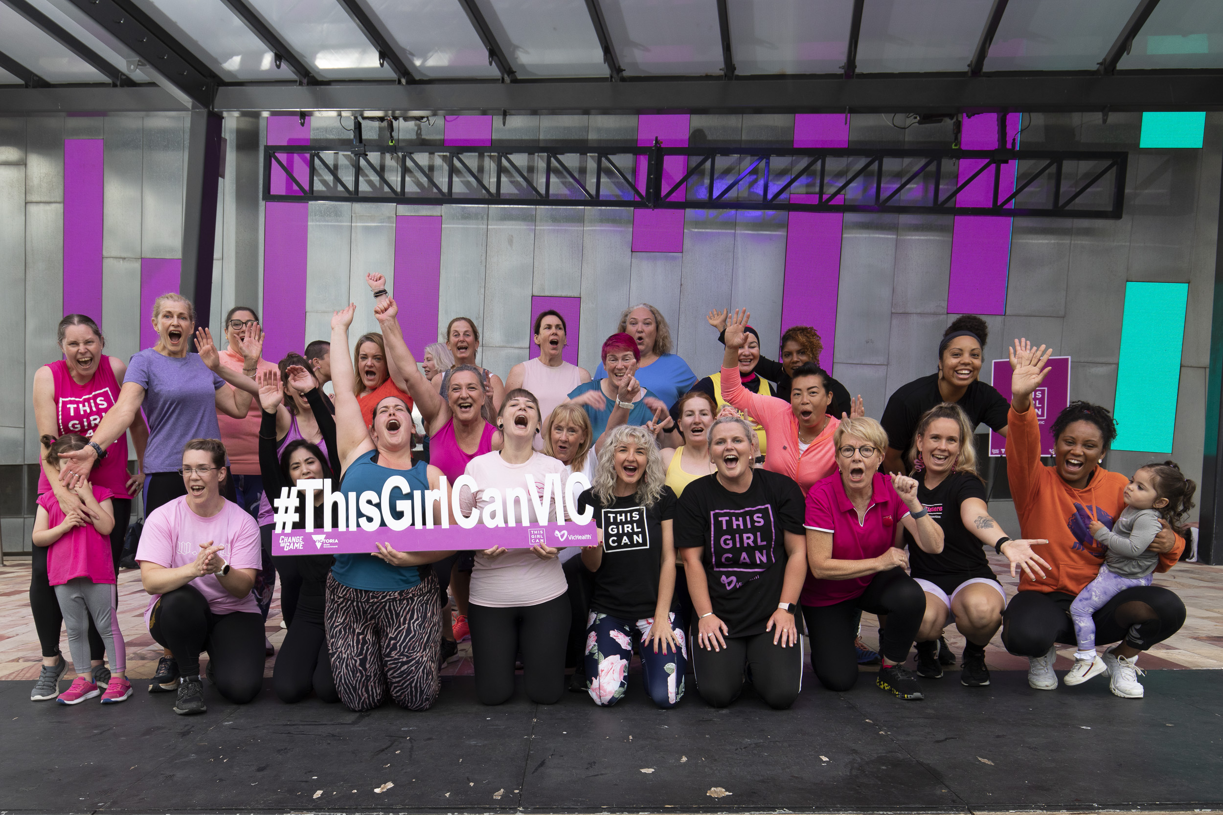 Women in sports gear and This Girl Can shirts on a stage holding a #ThisGirlCan sign