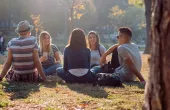Five young adults are sitting under a tree in the park. The sun is shinning and they are smiling at each other, looking relaxed. 