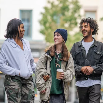 Three friends are walking down the street they are drinking coffee. One person looks like they've told a joke as the other two are laughing and smiling affectionately at them.