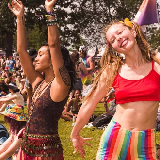 Two women, wearing festival attire, are dancing in the sunshine.