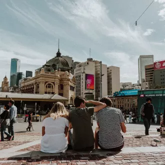 Three young adults sit facing Flinders Street Station. Thier backs are to camera.
