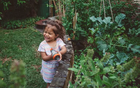 A photo of a child smiling as she waters a garden of fresh vegetables and herbs