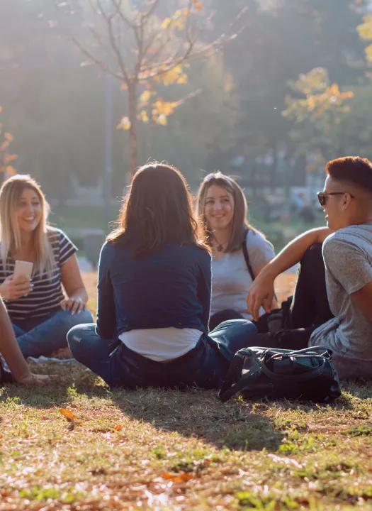 Five young adults are sitting under a tree in a park. The sun is shinning and they are all smiling at each other. 