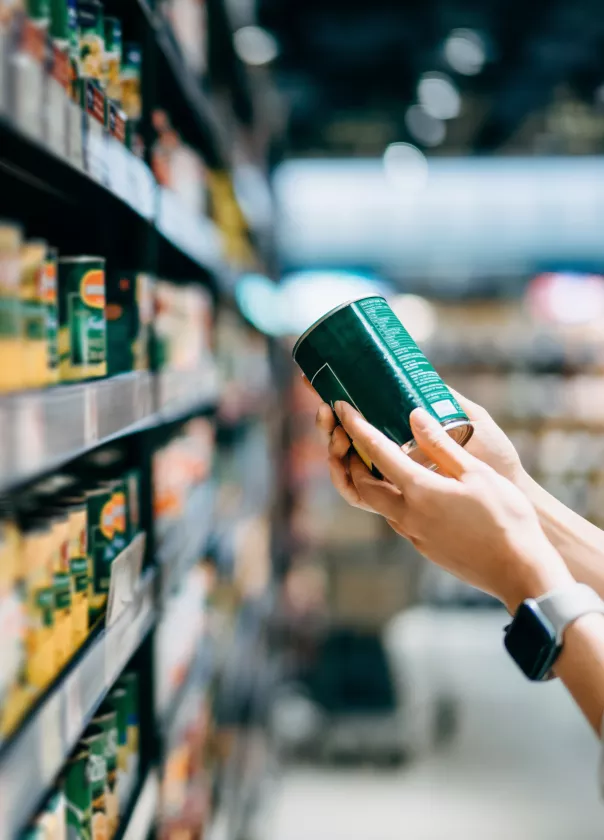 We see a supermarket shelf filled with tinned food. A hand is coming into the right hand side of the frame holding a can as if they are inspecting it. 