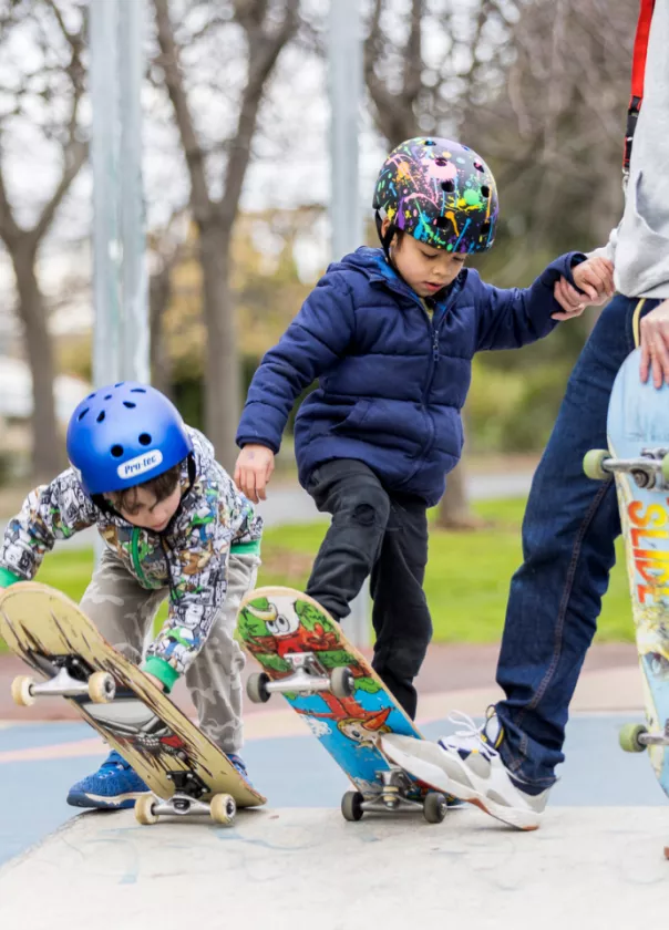 A photo of two young kids learning to skateboard wearing brightly coloured helmets.