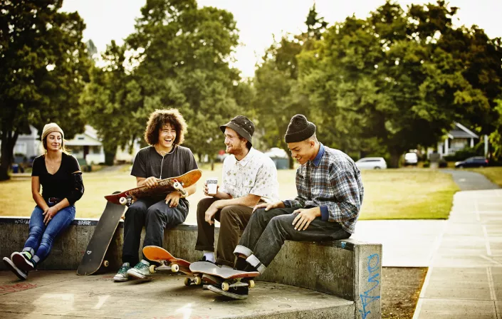 Four young adults are sitting on a wall, drinking coffee and holding their skateboards. The sun is shinning and they are all smiliing and laughing. 