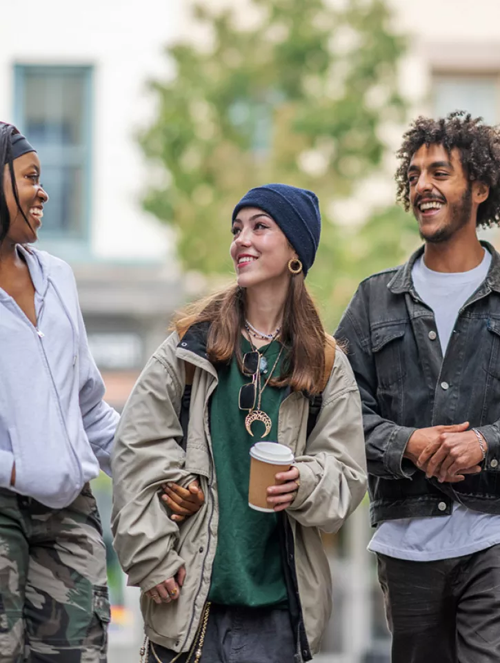 Three friends are walking down the street they are drinking coffee. One person looks like they've told a joke as the other two are laughing and smiling affectionately at them.