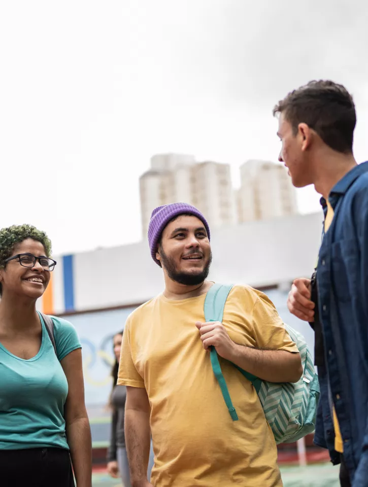 Three young adults are standing on a basketball court. They have backpacks on and look happy, like they are making plans to do something fun. 