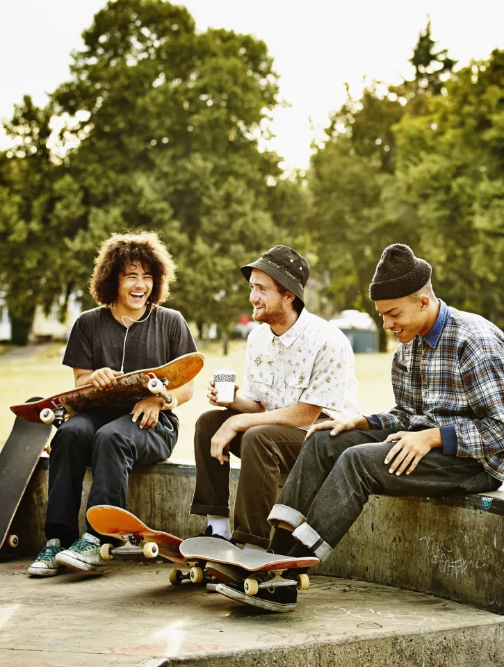 Four young adults are sitting on a wall holding their skateboards. They are drinking coffee and laughing.