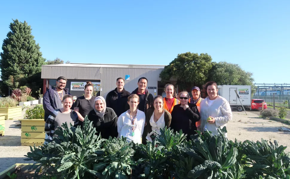 a group of people are standing in front of a garden bed flourishing with kale . They are all posing for the camera.